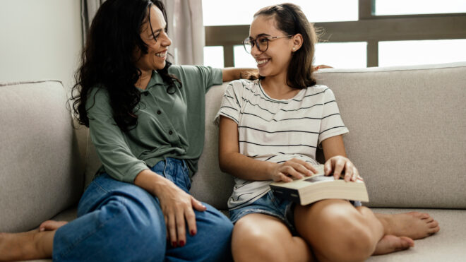 A mother and her teenage daughter laughing happily on the sofa after a satisfying read, the girl holding the book on her leg.