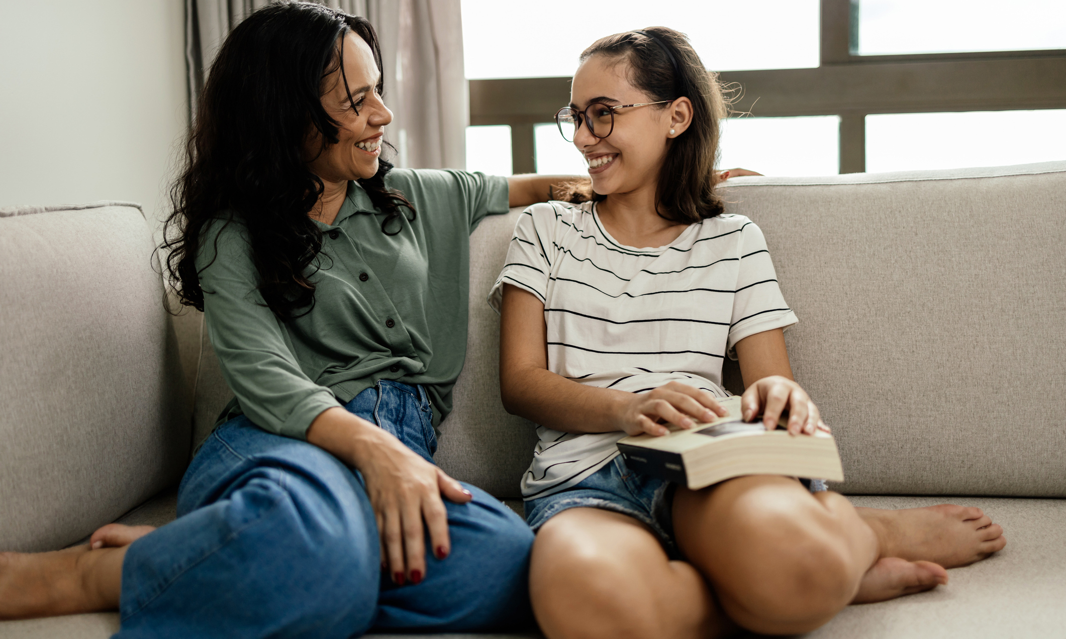 A mother and her teenage daughter laughing happily on the sofa after a satisfying read, the girl holding the book on her leg.