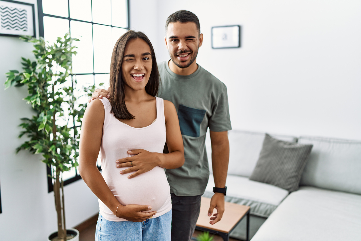 Young interracial couple expecting a baby, touching pregnant belly winking looking at the camera with sexy expression, cheerful and happy face.