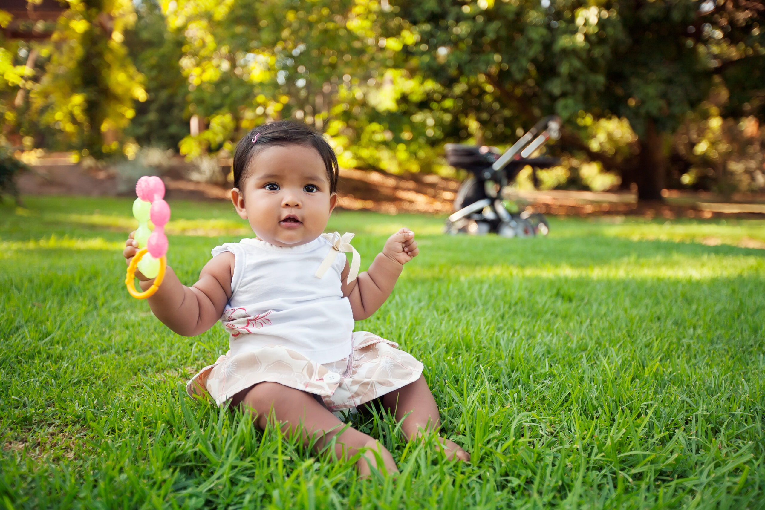 A happy hispanic baby playing and grasping a few chewable baby toys while sitting on the lawn of a park with he stroller in the background.