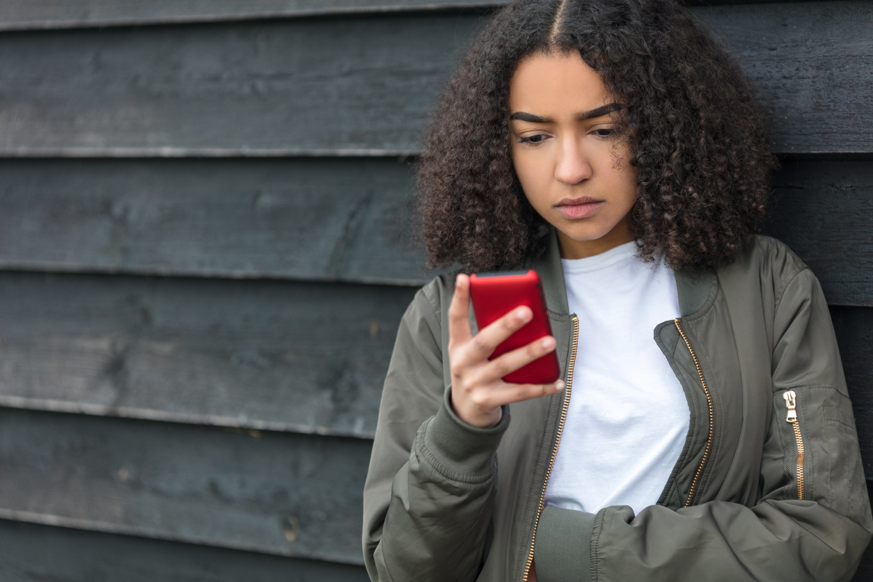 Outdoor portrait of beautiful sad depressed mixed race African American girl teenager female young woman texting on red cell phone wearing green bomber jacket