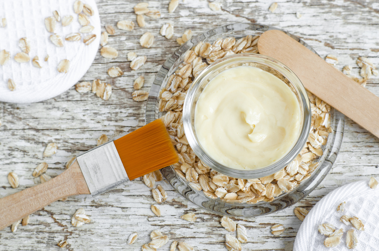 Small glass bowl with facial mask (moisturizer), body butter with oat extract. Natural cosmetics ingredients. Wooden background. Top view, copy space.