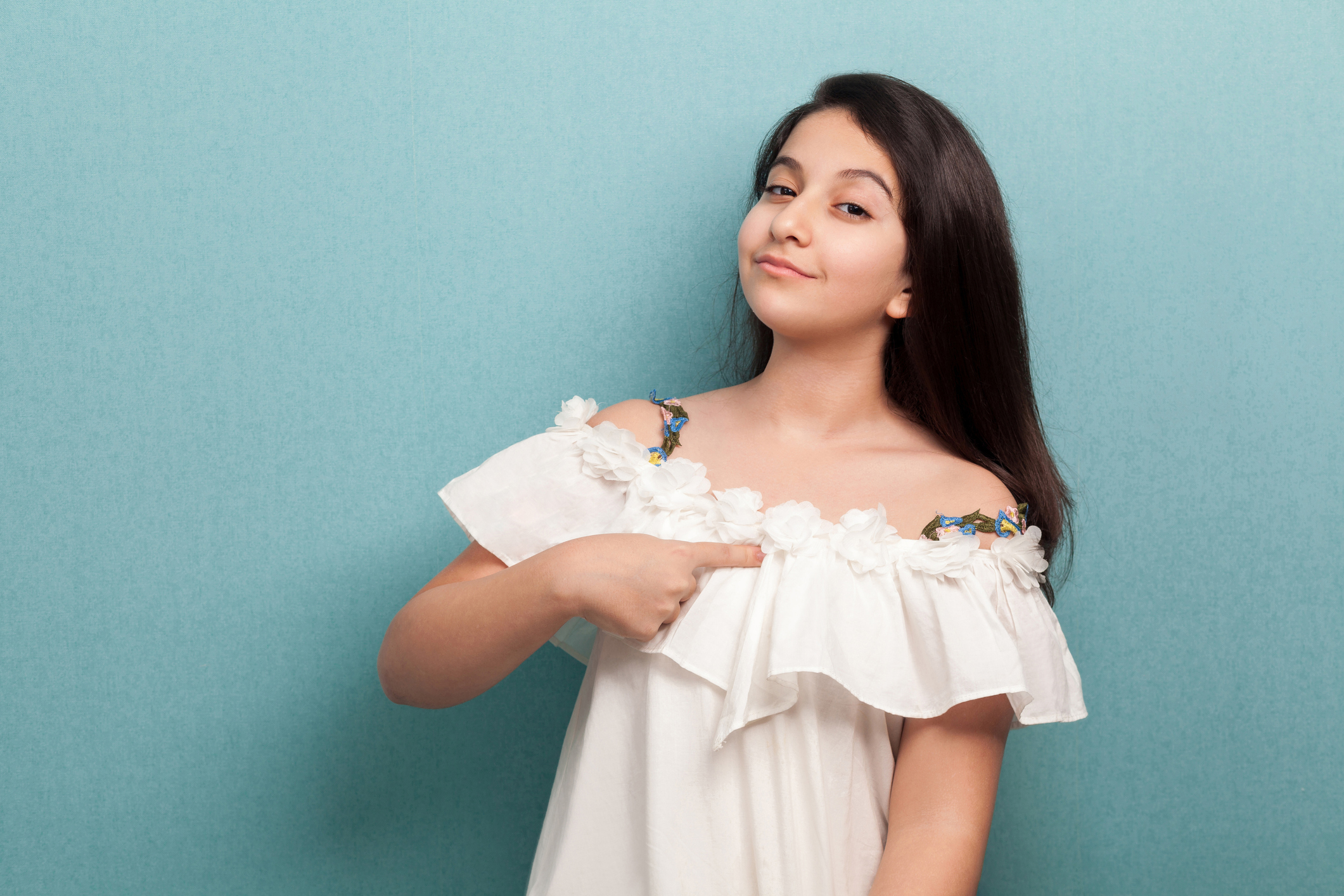 This is me. Portrait of proud beautiful brunette young girl with black straight hair in white dress standing, pointing herself and looking at camera.