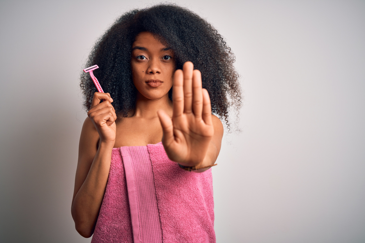 Young african american woman with afro hair in a towel holding female razor for body depilation with open hand doing stop sign with serious and confident expression, defense gesture