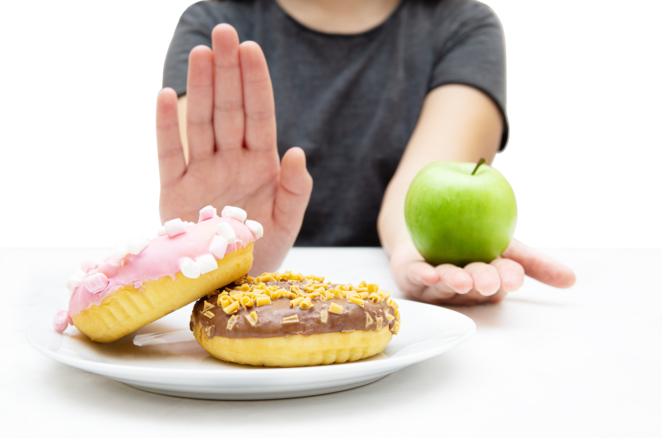 Woman choosing a fresh apple over a plate of donuts, rejecting junk unhealthy food with a hand gesture. Closeup, selective focus.