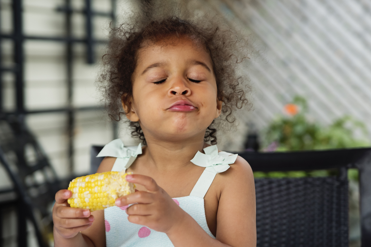 Mixed-race toddler eating corn outdoors in summer.