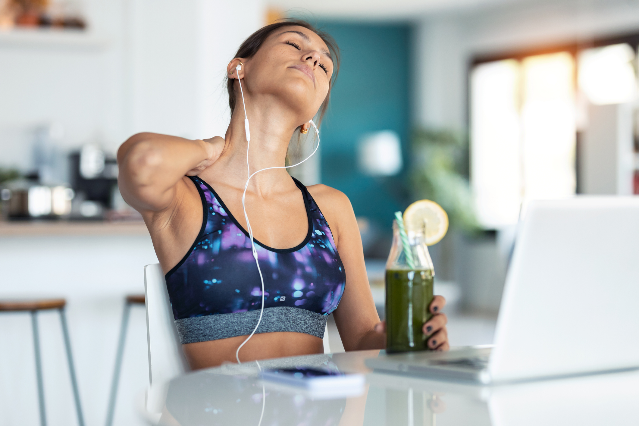 Tired young sporty woman suffering neck pain while drinking detox juice in the kitchen at home.