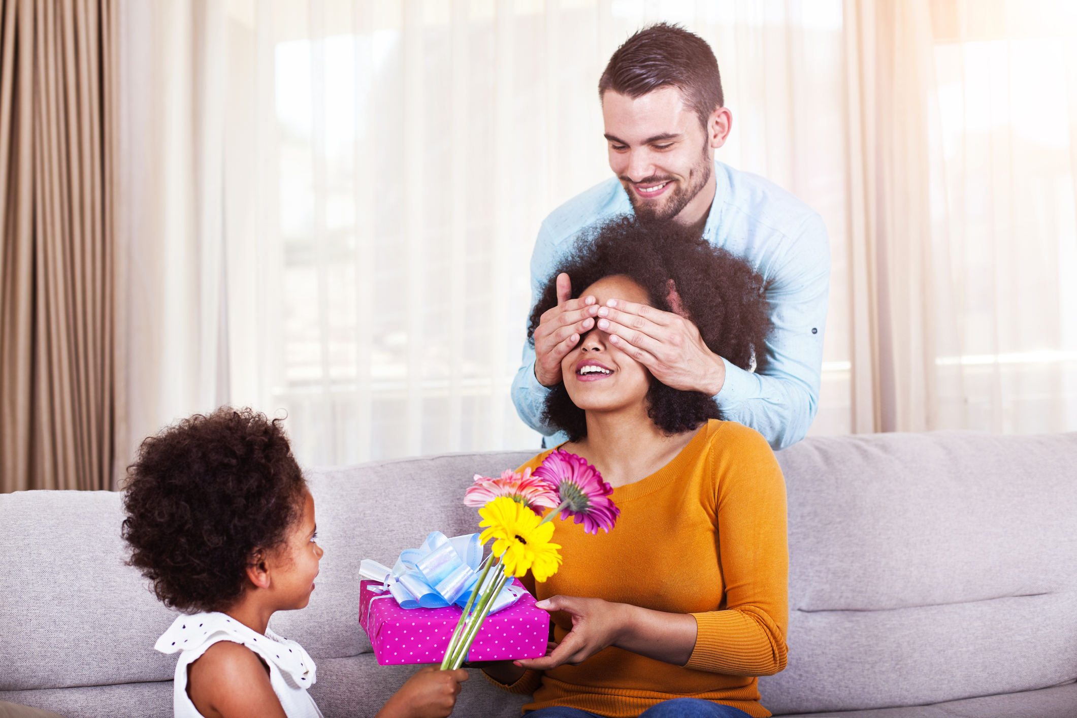 Father holding gift and flowers, daughter covering mothers eyes