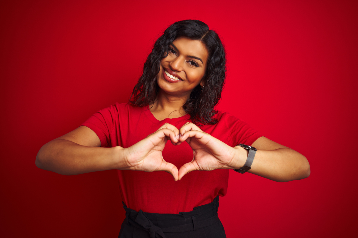 Beautiful transsexual transgender woman wearing t-shirt over isolated red background smiling in love doing heart symbol shape with hands. Romantic concept.