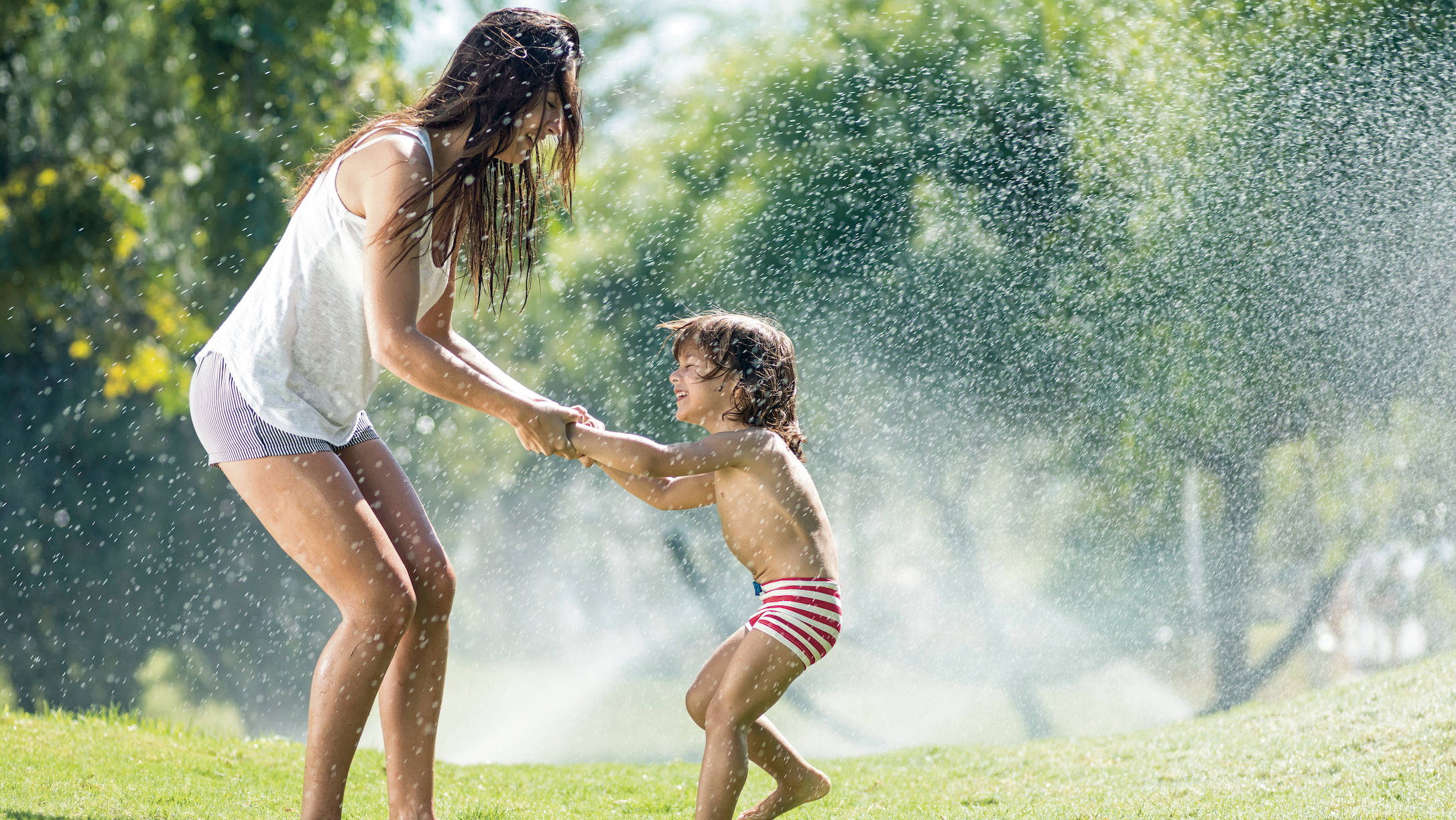 Mother And Children playing Through Garden Sprinkler