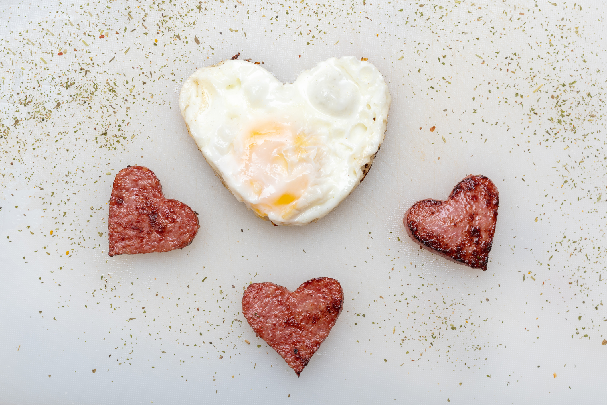 Heart shaped scrambled eggs with heart shaped sausages, herbs and spices on a white plate, top view