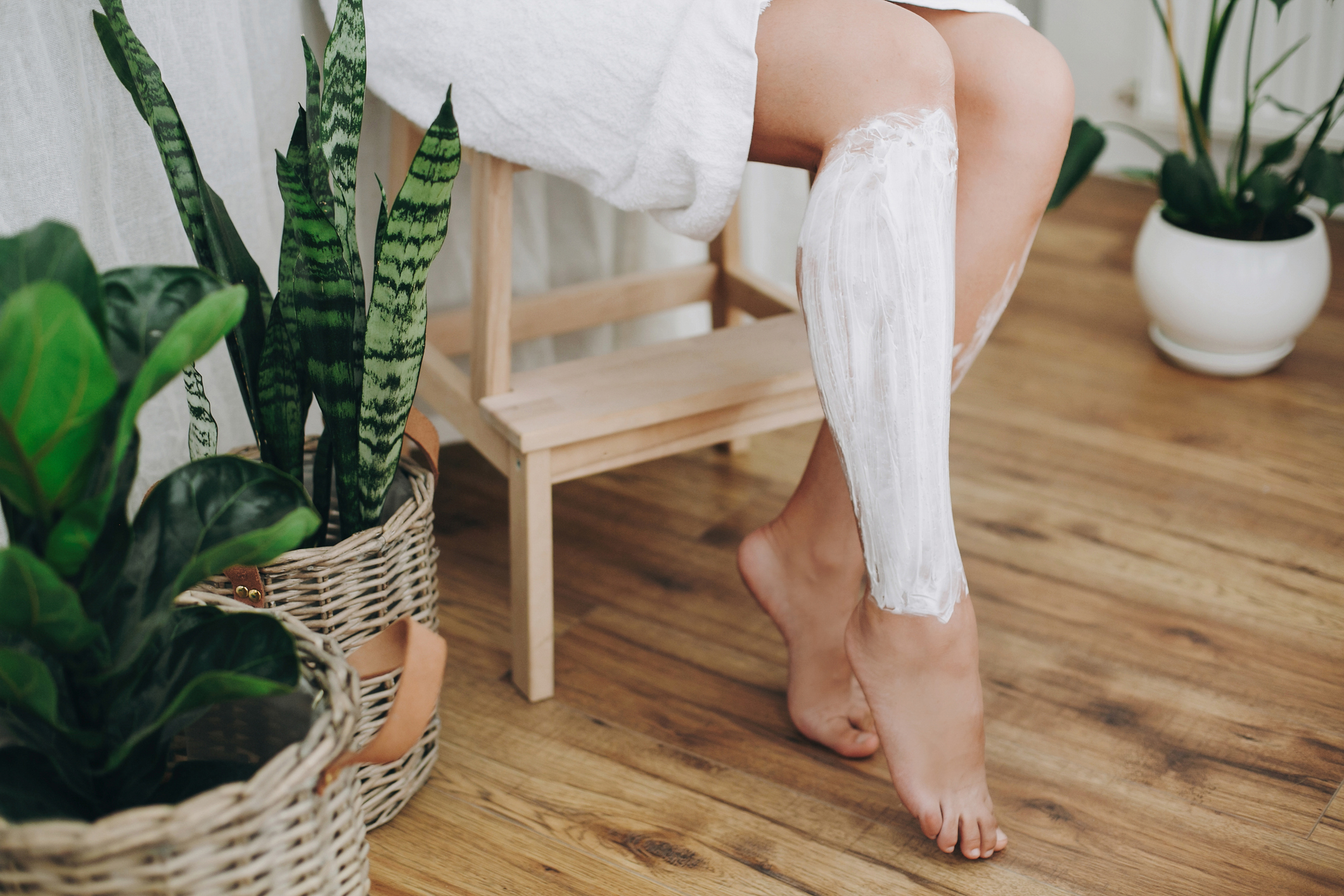 Hair Removal concept, depilation cream. Young woman in white towel applying shaving cream on her legs in home bathroom with green plants. Skin care and wellness concept.