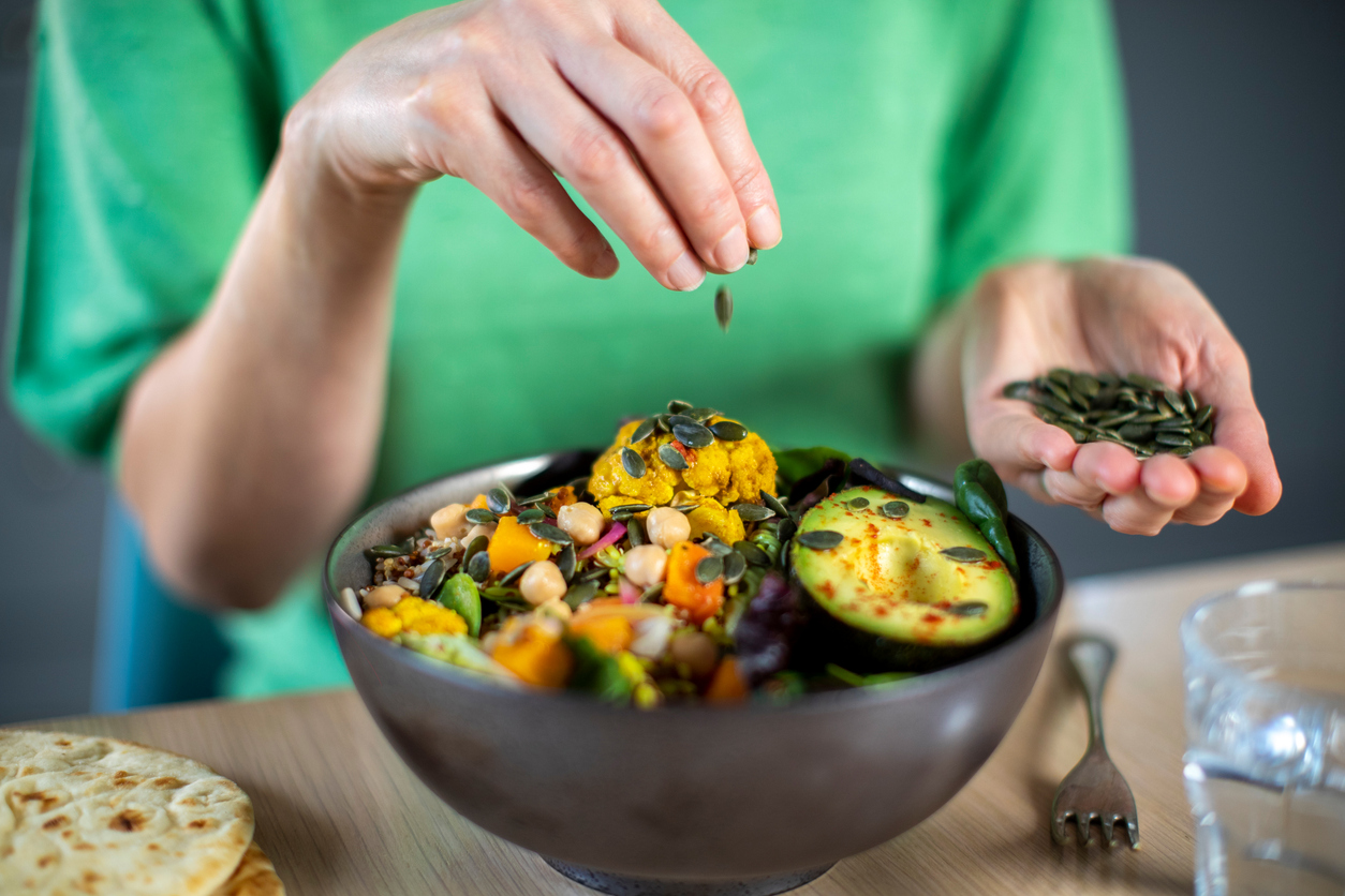 Close Up Of Woman Adding Pumpkin Seeds To Healthy Vegan Meal In Bowl