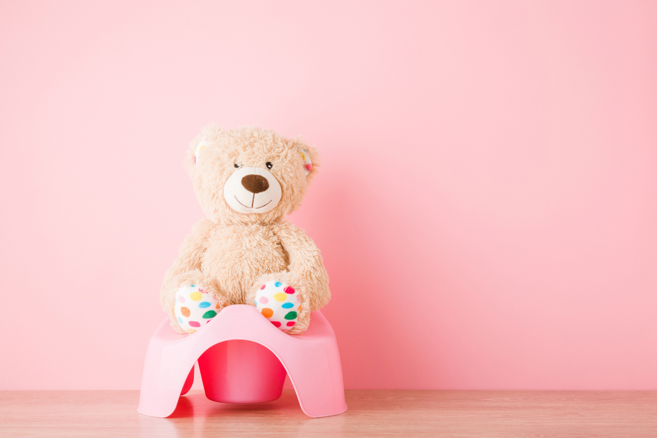 Brown teddy bear sitting on baby potty on floor. Front view. Closeup. Empty place for text on light pink background. Pastel color.