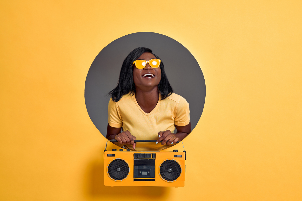 Image of beautiful young woman in yellow sunglasses laughing and holding vintage boombox isolated over circle hole in yellow background. Copy space.