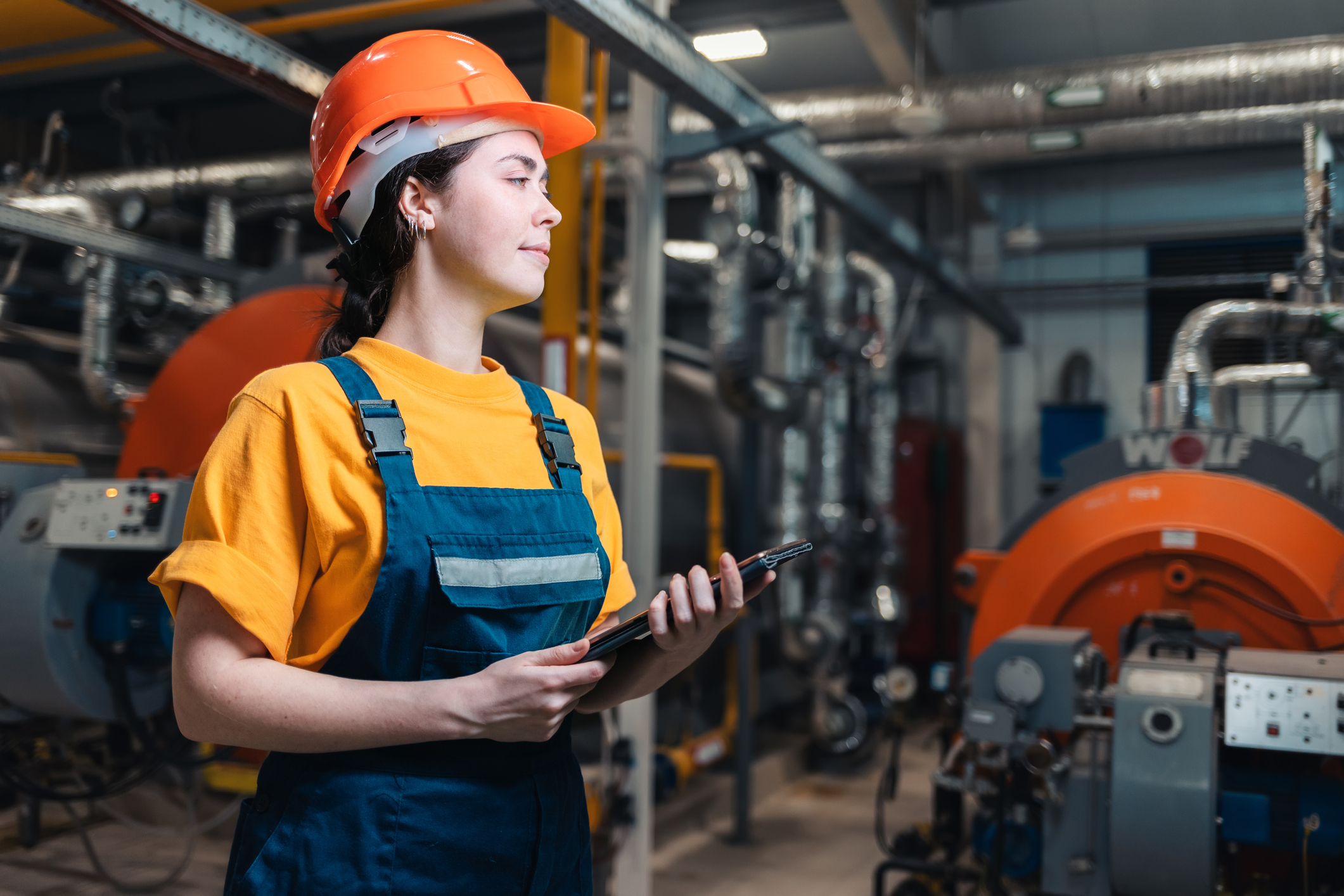 Portrait of a caucasian female worker in uniform and helmet with a tablet in her hand. In the background boiler room. Concept of industrial production and equality. Side view