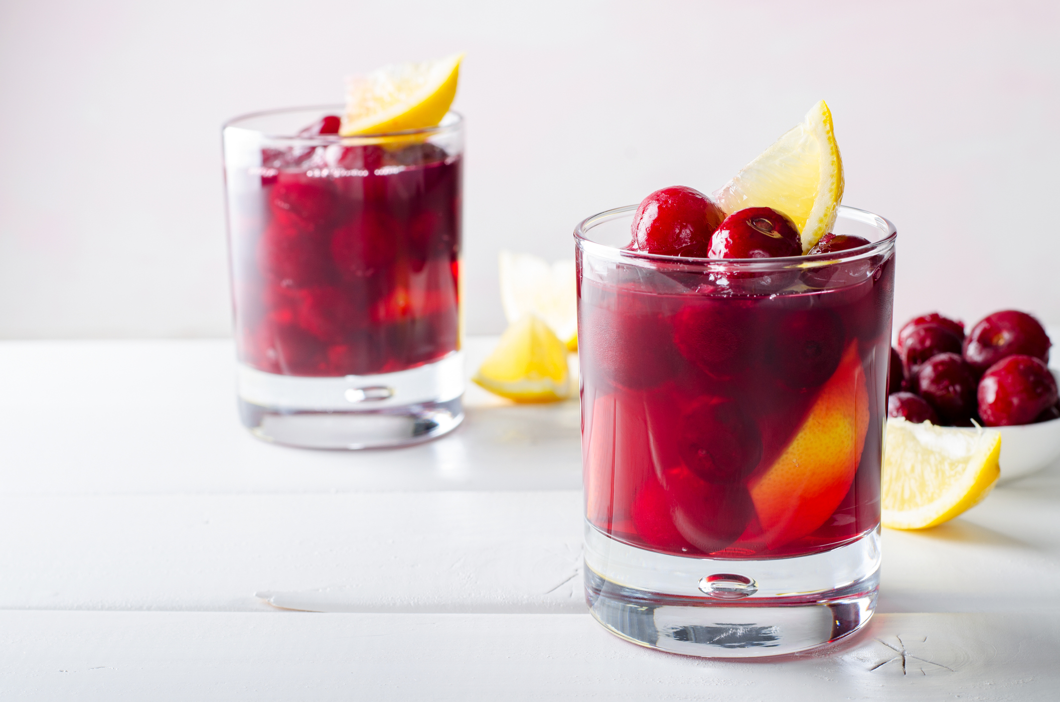 Cold drink with cherry and lemon in glasses, on white wooden background