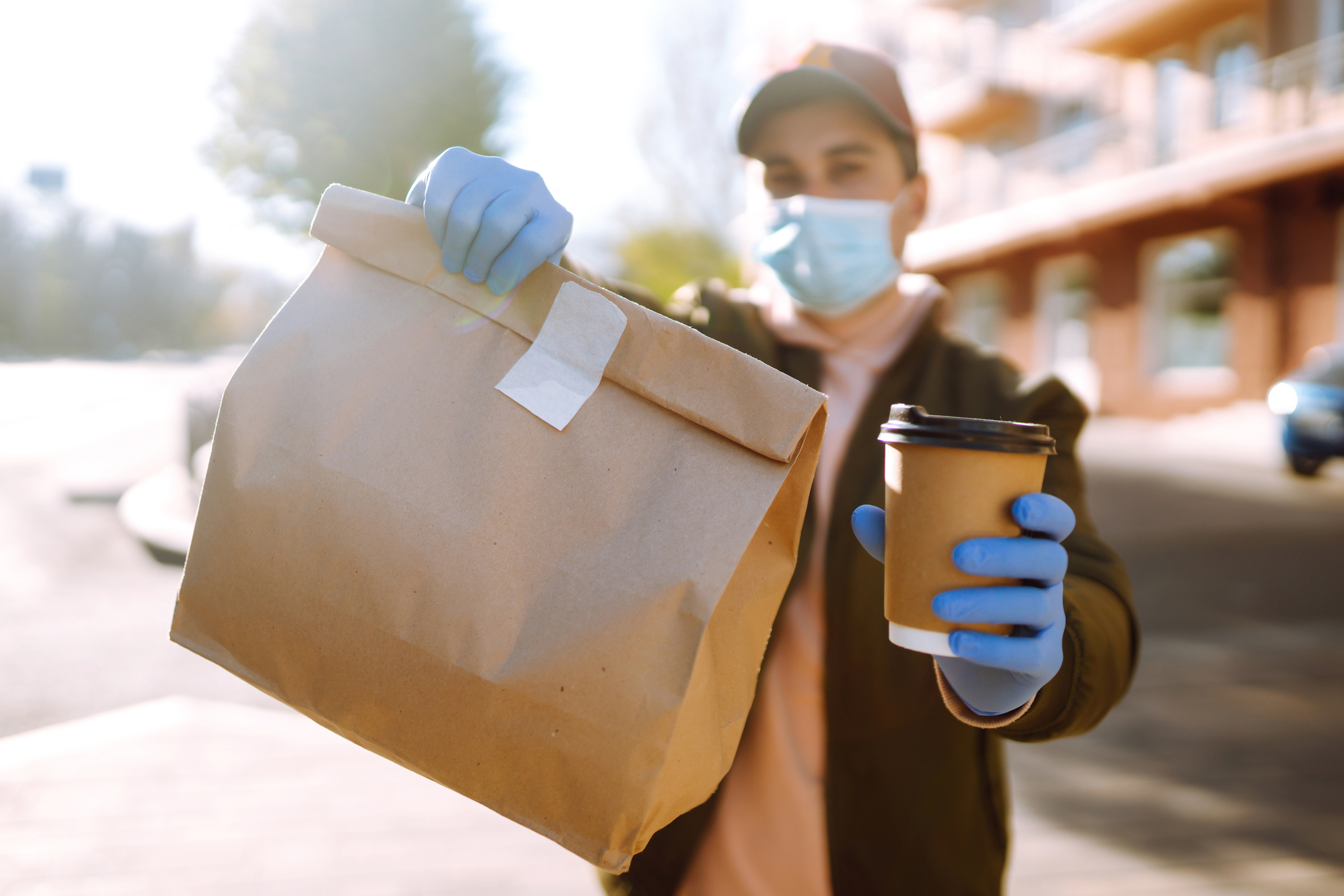 Courier in protective mask and medical gloves holds out paper packet with food and cofee.