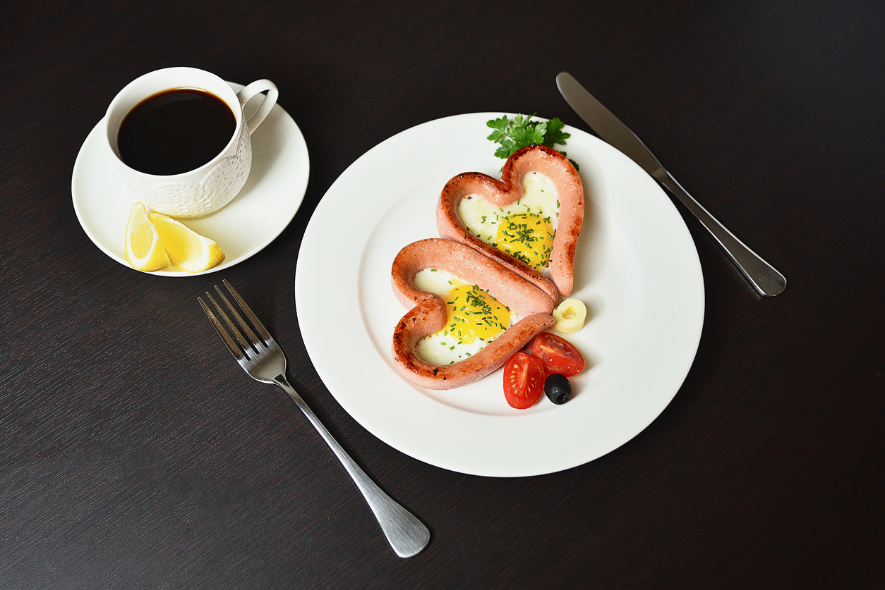 Scrambled eggs in the form of heart on a white plate with sausages, tomatoes, greens and coffee with lemon