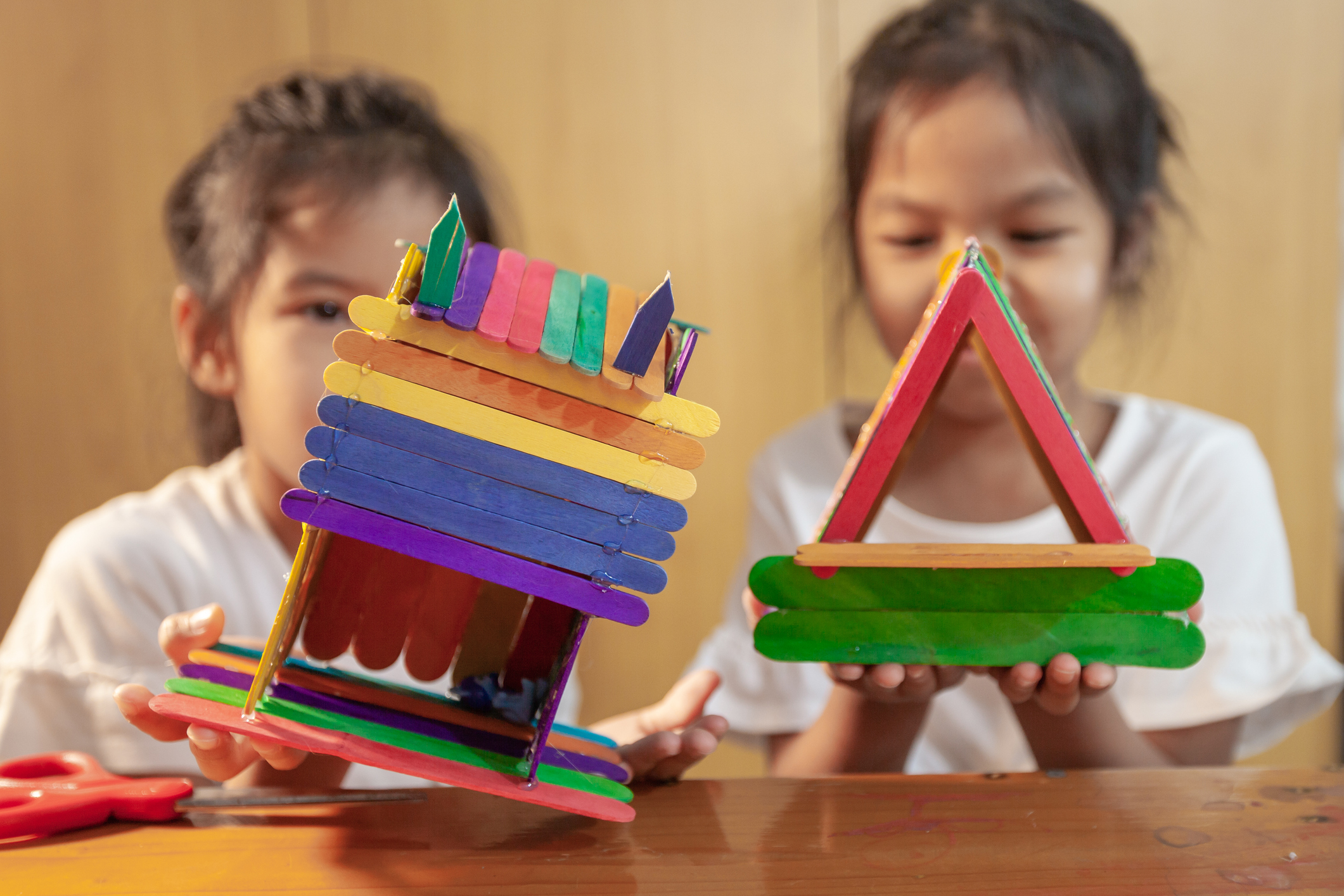 Asian child girl glueing colored ice cream sticks by hot melt electrical glue gun. Children have fun to make house on a handicraft project.