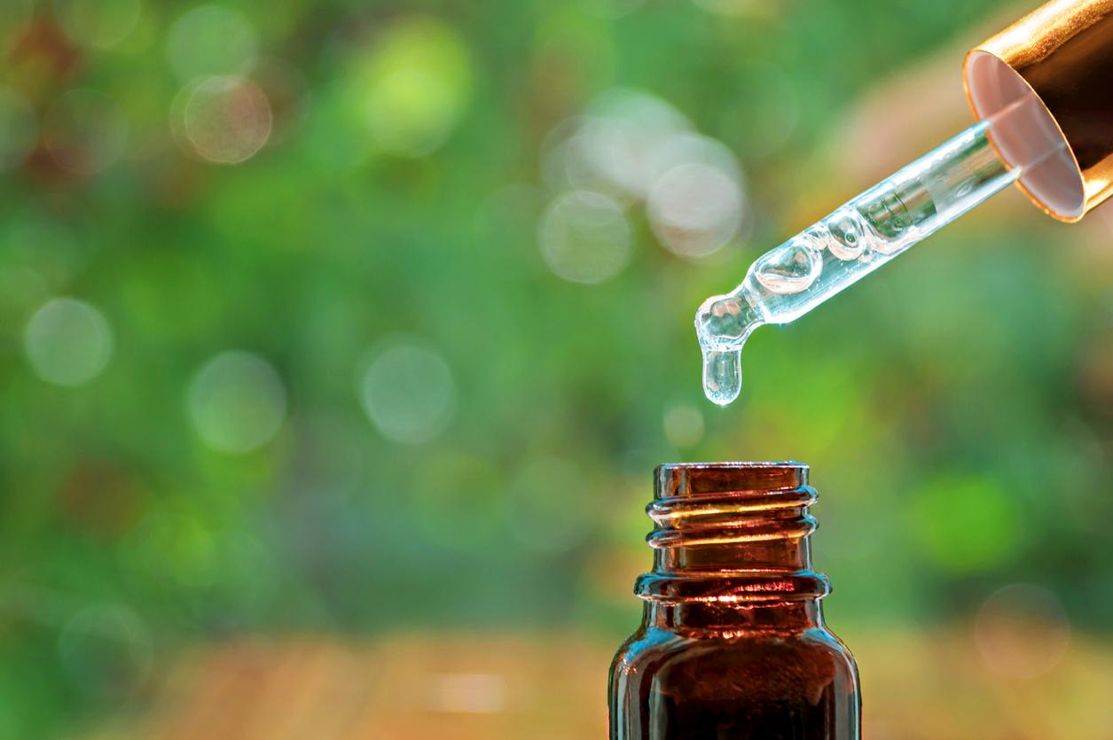 Pipette with essence drop and bottle, closeup on blurred nature background. Herbal essential massage oil dripping into bottle. Spa beauty concept. Selective focus