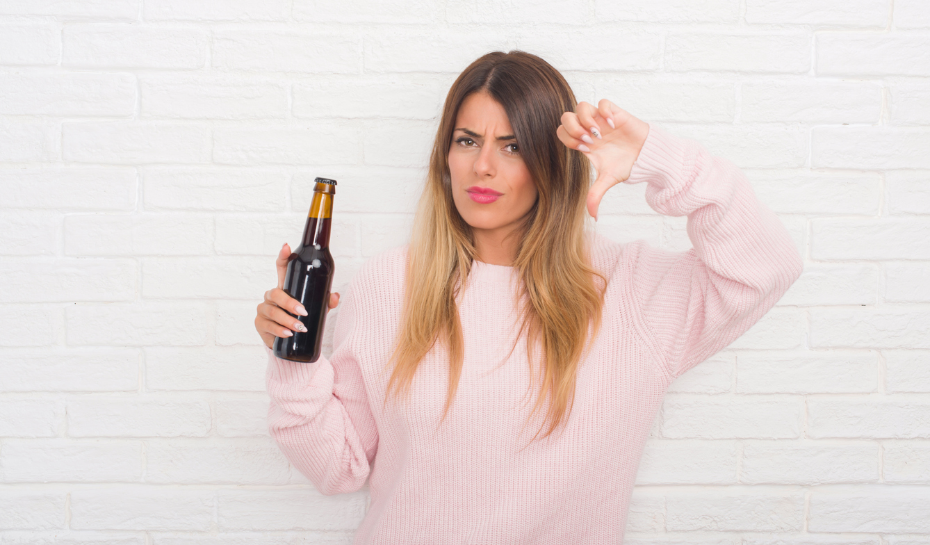 Yound adult woman over white brick wall holding beer with angry face, negative sign showing dislike with thumbs down, rejection concept