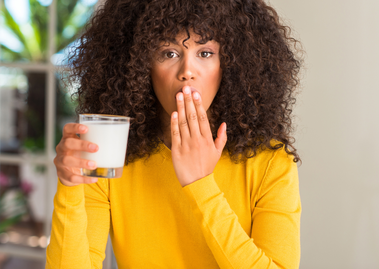 African american woman holding a glass of milk cover mouth with hand shocked with shame for mistake, expression of fear, scared in silence, secret concept