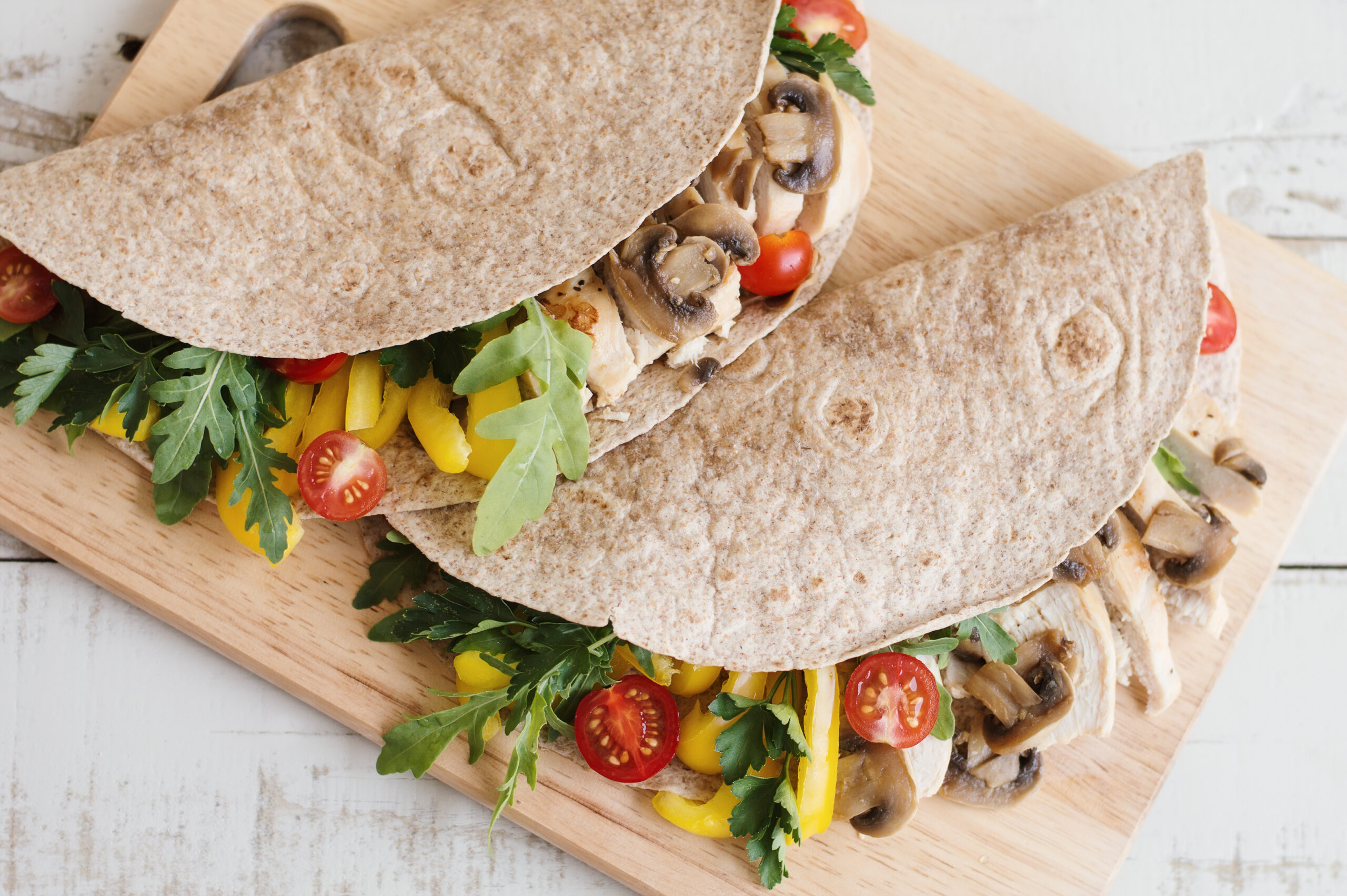 Two sandwiches with whole wheat wrap, chicken breast, mushroom and seasonal vegetables served on wooden board. Healthy and balanced meal