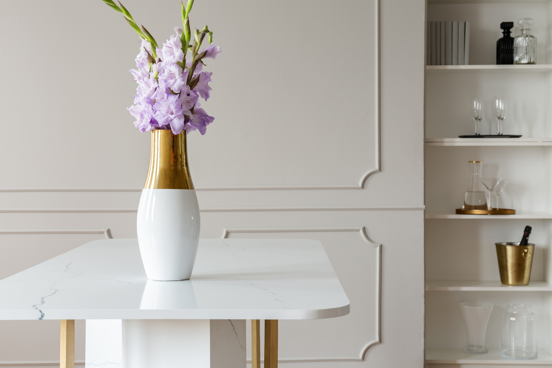 Lavender purple flowers in a golden vase on a white marble table in an elegant dining room interior with molding on beige walls