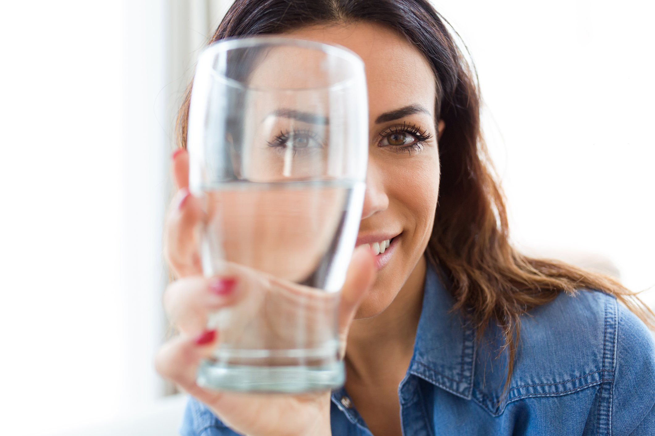 Pretty young woman smiling while looking at the camera through the glass of water at home.