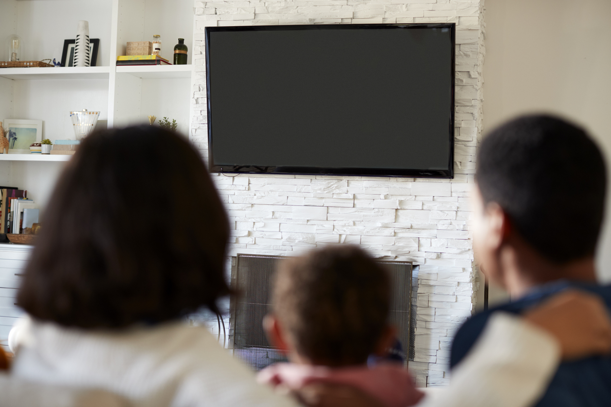 Back view of young family sitting on the sofa and watching TV together in their living room, close up, focus on television