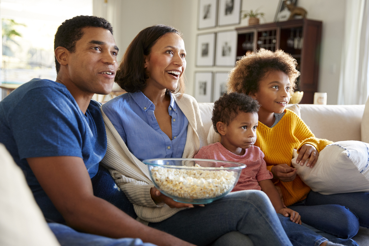 Young family sitting together on the sofa in their living room watching TV and eating popcorn, side view