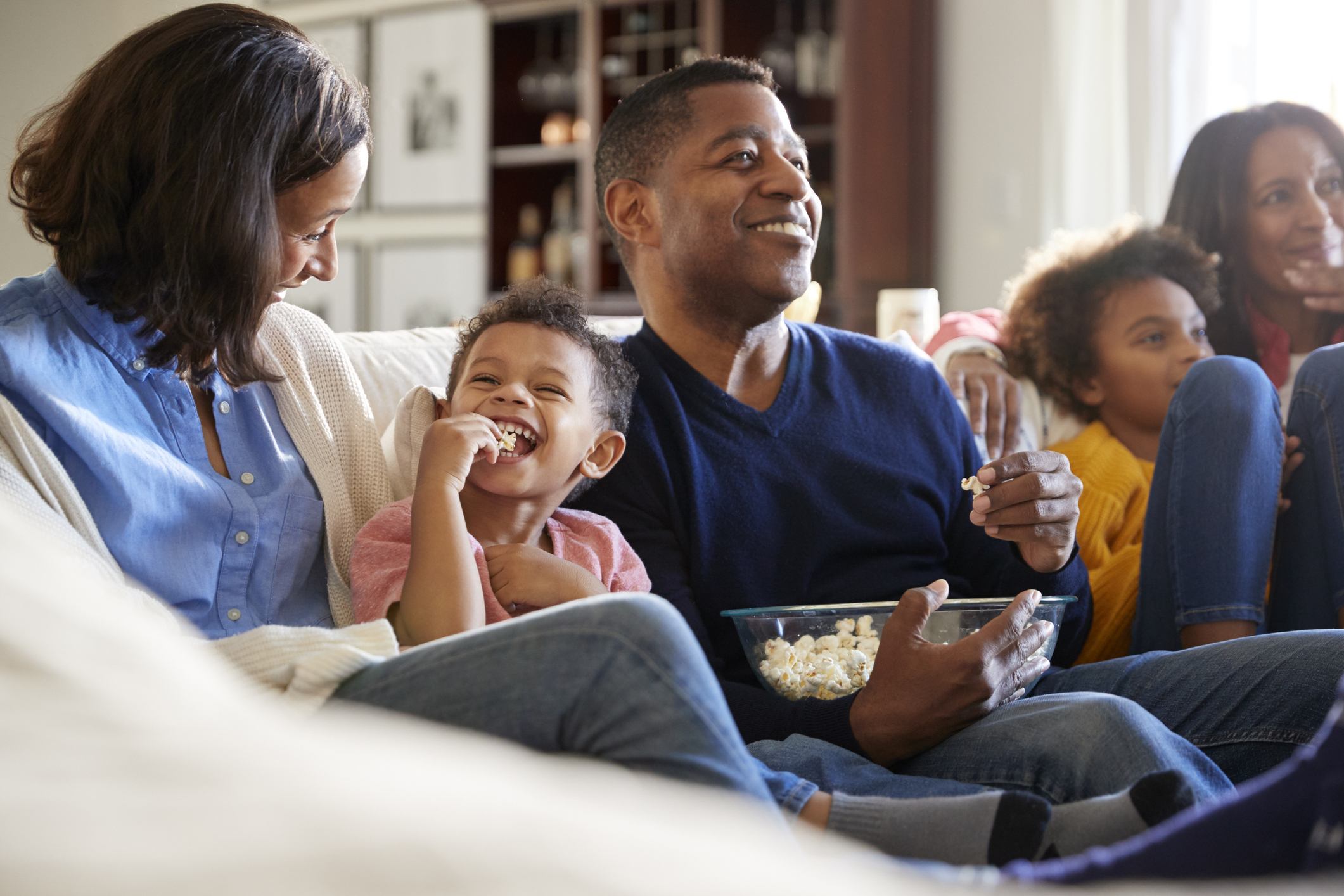 Three generation family family sitting on the sofa in living room, watching TV and eating popcorn, selective focus