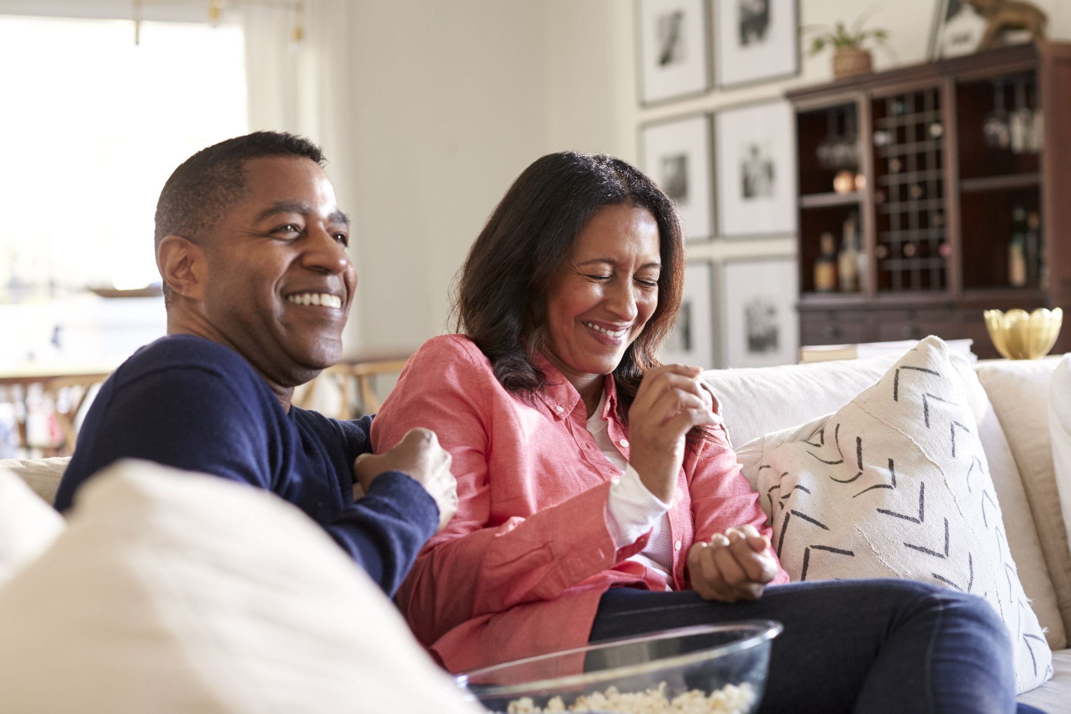 Close up of middle aged couple sitting on the sofa in their living room watching TV, laughing and eating popcorn, close up