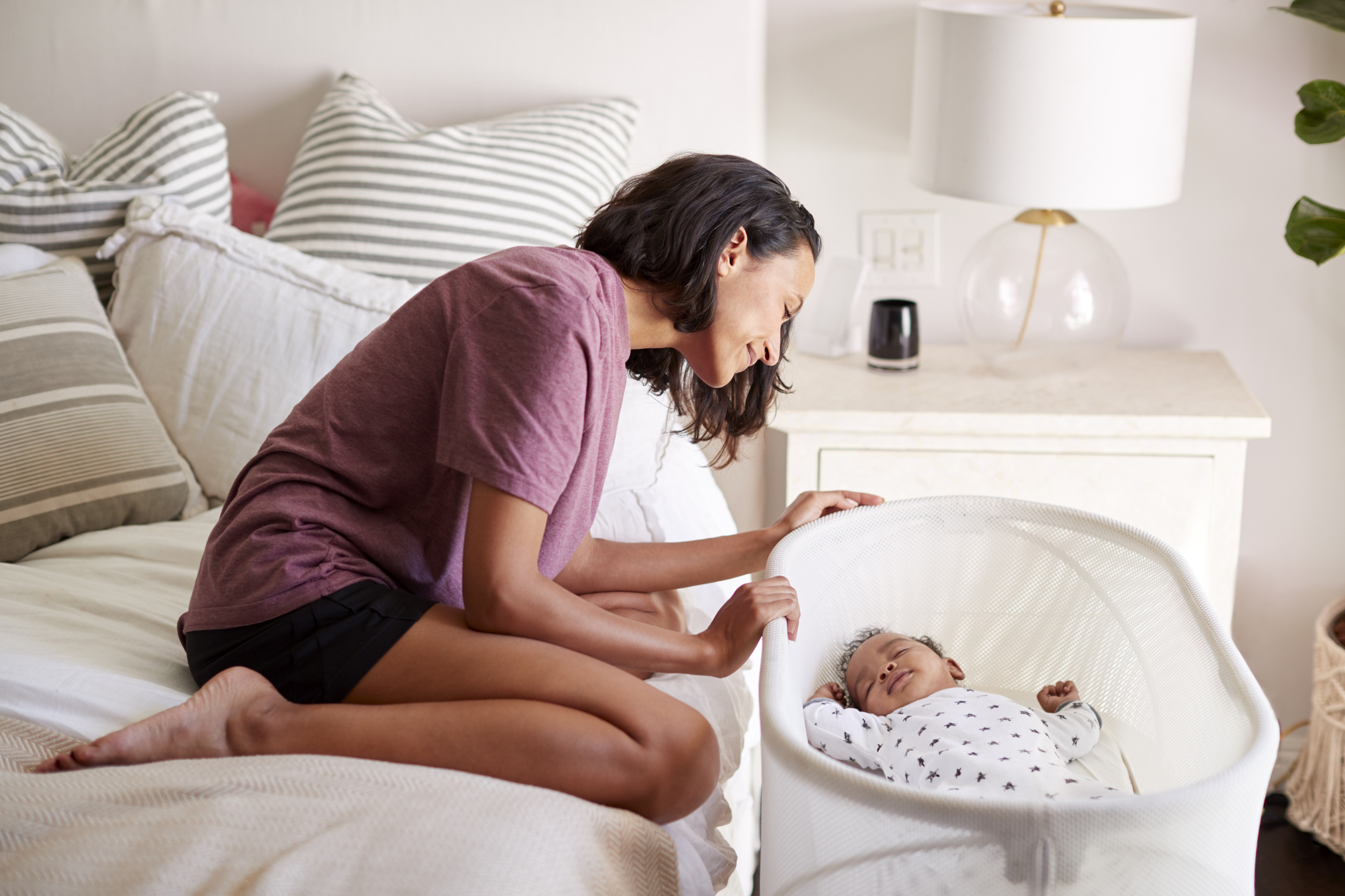 Young adult mother kneeling on her bed looking down at her three month old baby sleeping in his cot, side view