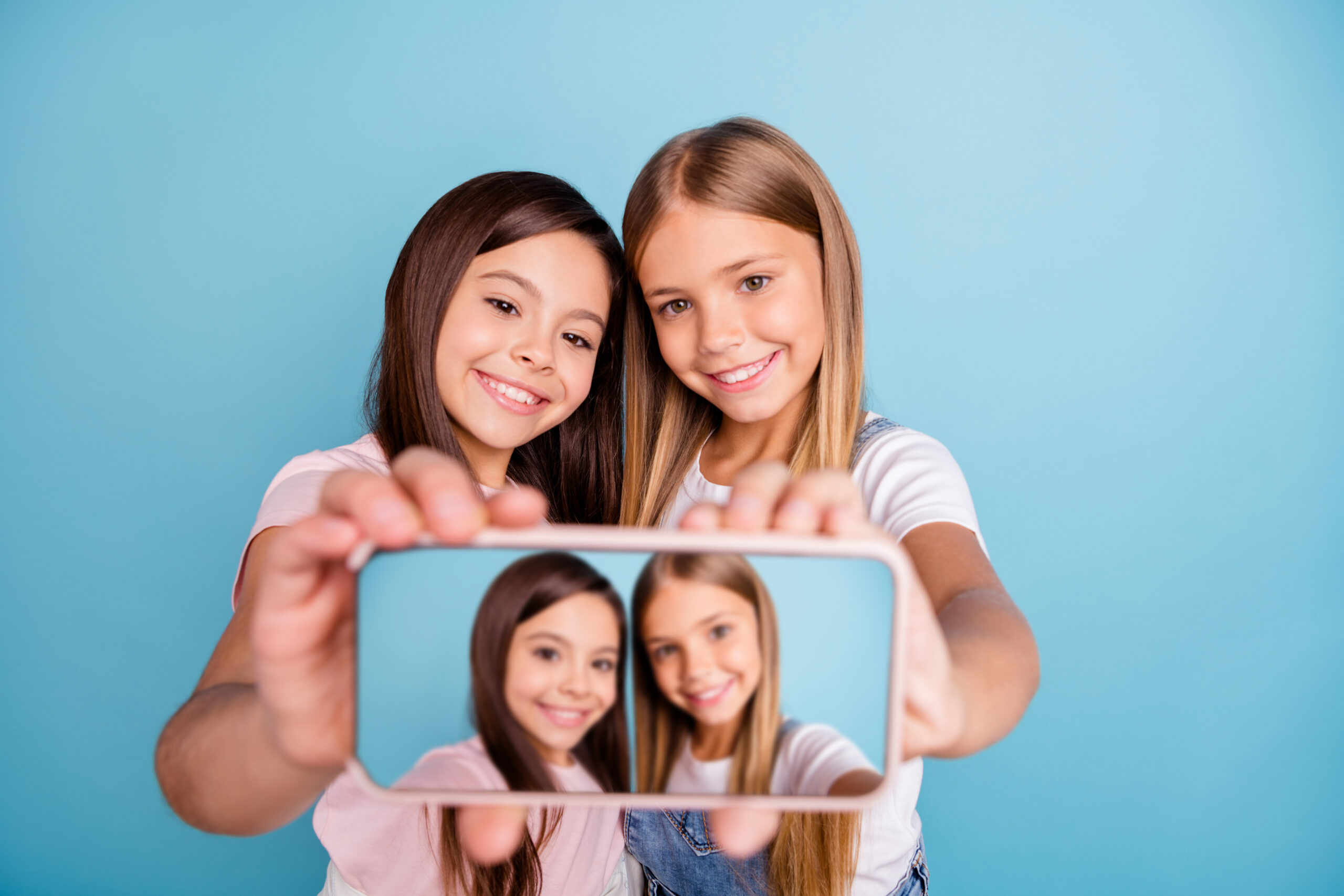 Close up photo two little she her blond brunette girls long pretty hair telephone make take selfies talk tell speak millennials wearing casual jeans denim t-shirts isolated blue bright background