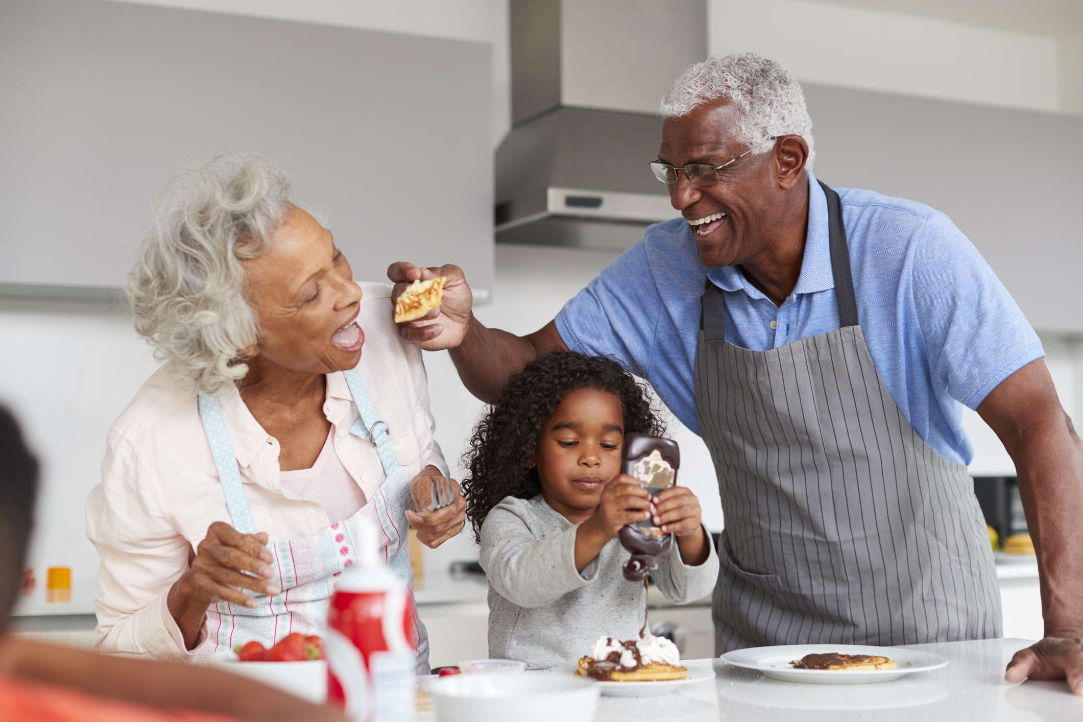 Grandparents In Kitchen With Granddaughter Making Pancakes Together