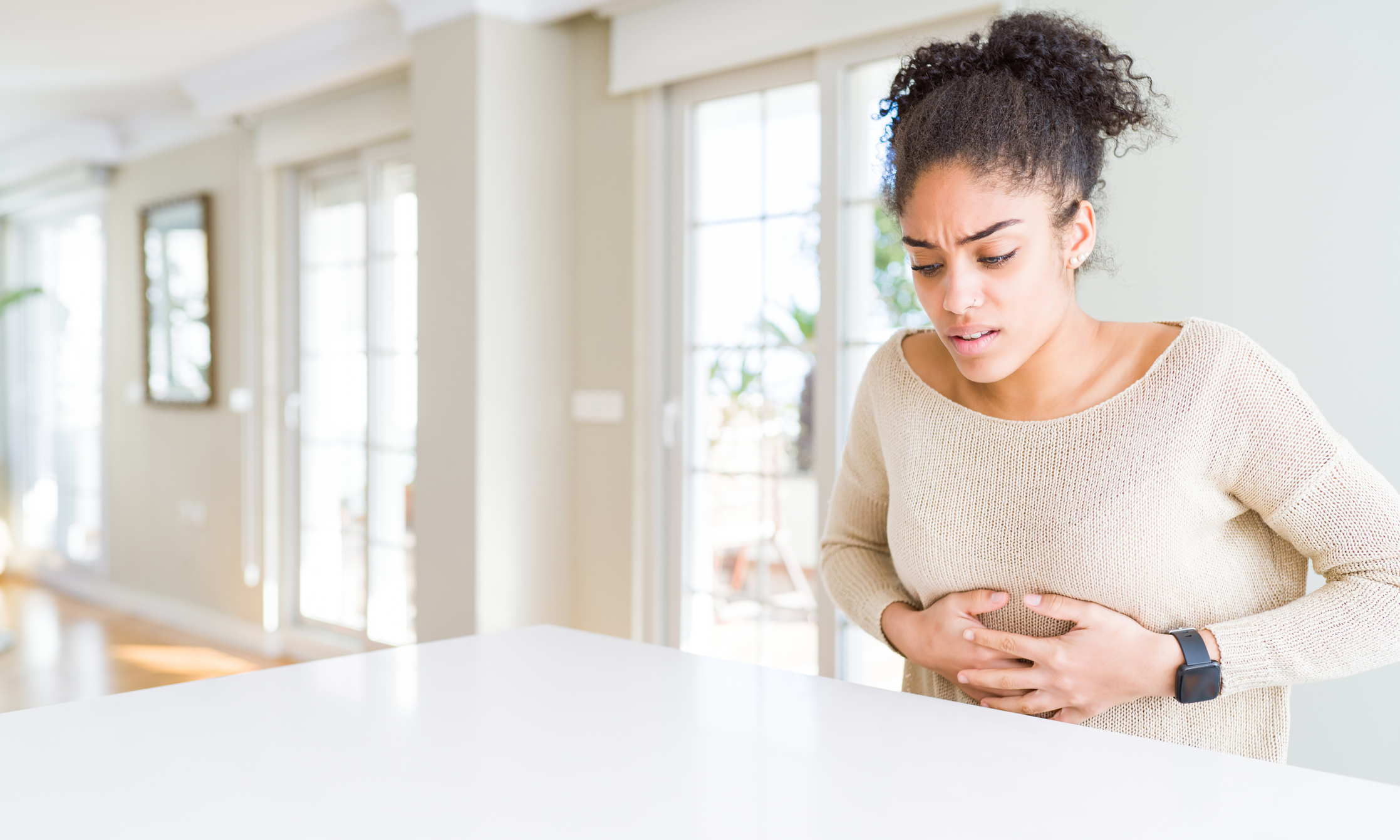 Beautiful young african american woman with afro hair sitting on table at home with hand on stomach because indigestion, painful illness feeling unwell. Ache concept.