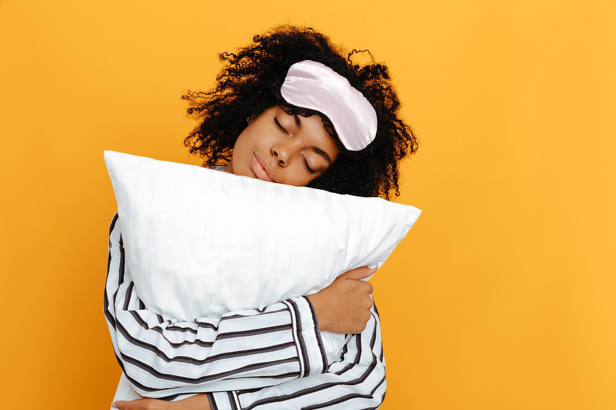 Sleeping. Dreams. Woman portrait. Afro American girl in pajama is hugging a pillow, on a yellow background