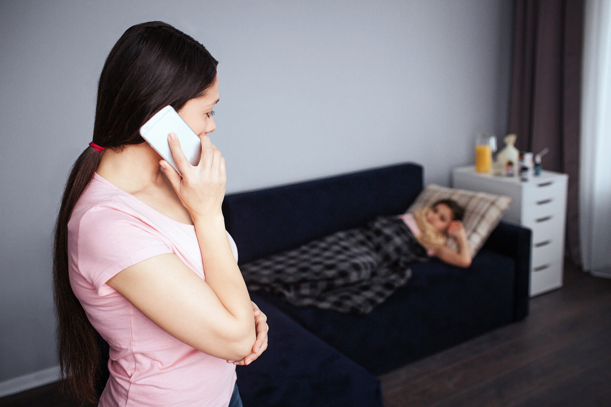 Young brunette stand in room and talk on phone. She look at her sick daughter. Girl lying on couch. She covered with blanket.