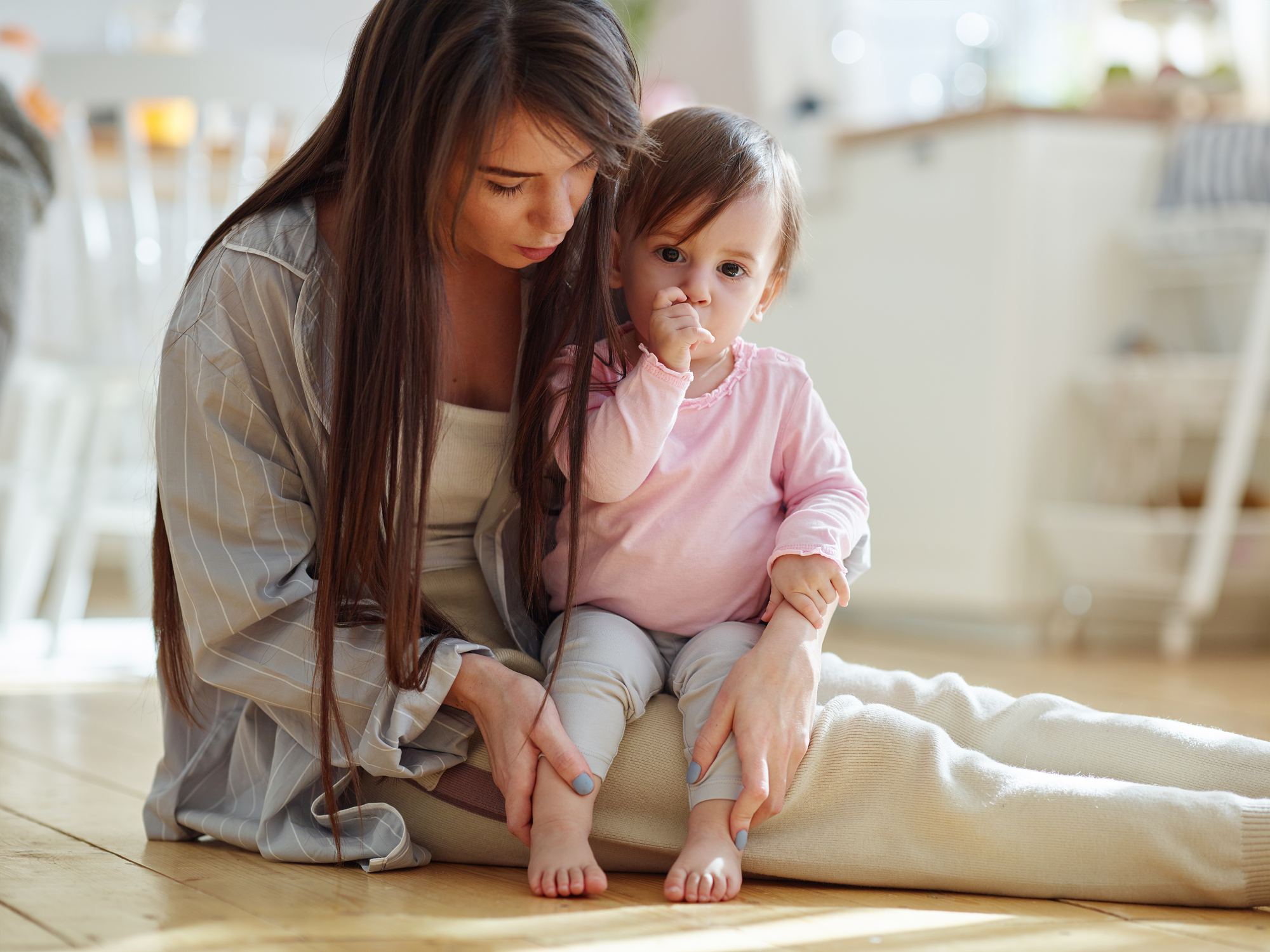 Loving young mother sitting on wooden floor at home and stroking legs of adorable baby daughter sitting on her lap, sucking thumb and looking at camera