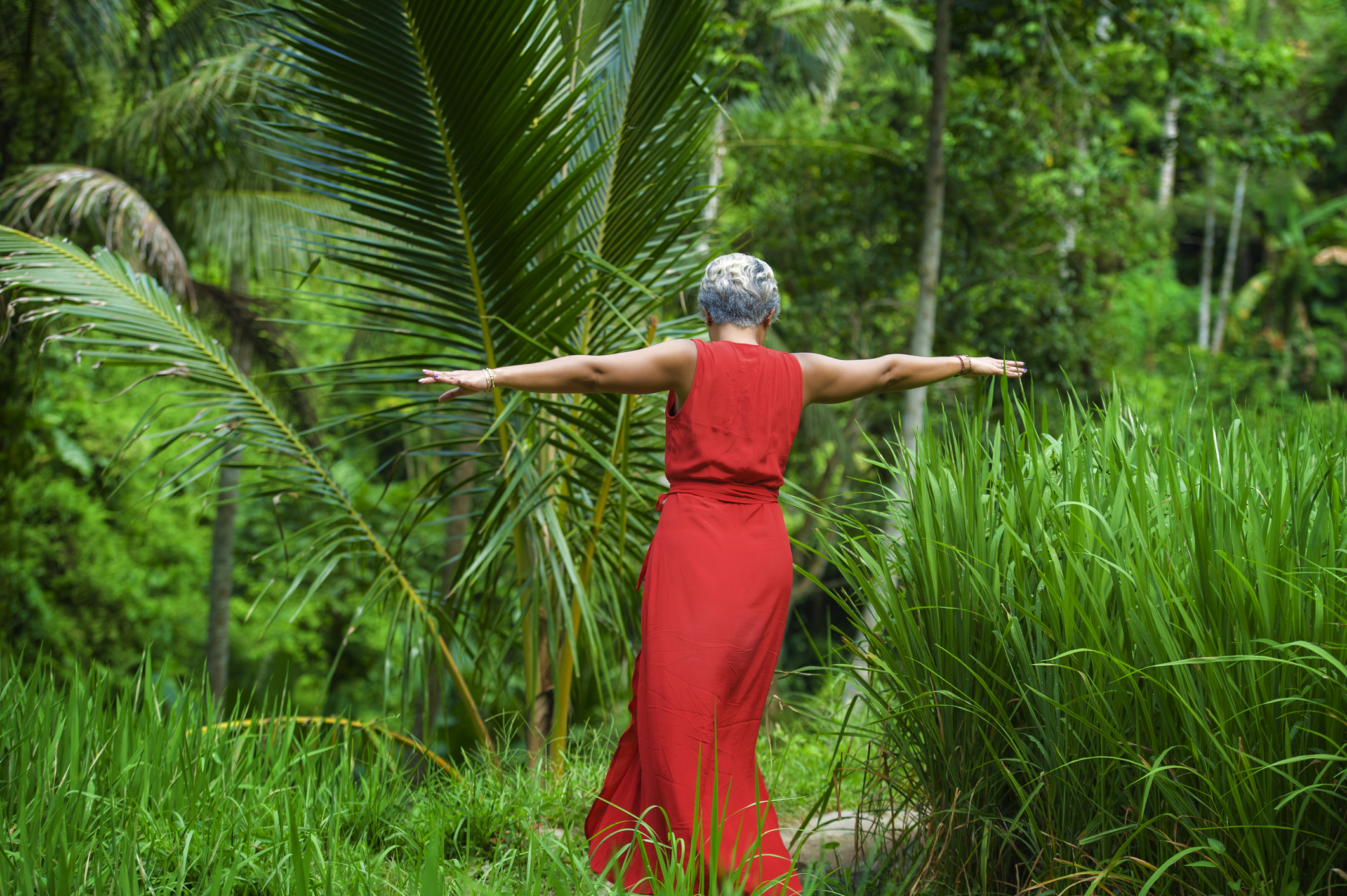 natural lifestyle portrait of carefree and happy middle aged 40s or 50s woman with grey hair in stylish red dress walking on green tropical forest landscape enjoying adventure holidays trip