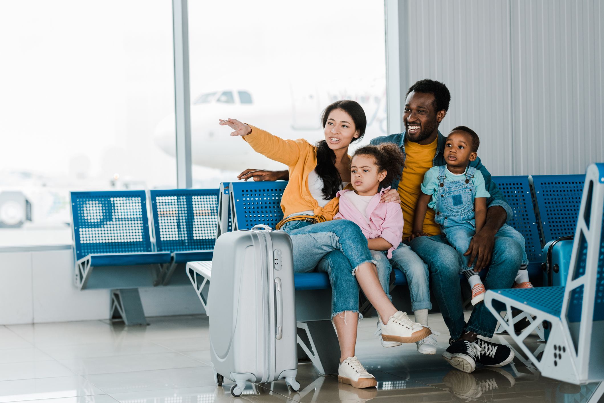 smiling african american family with baggage and kids sitting in airport while mother pointing with finger away