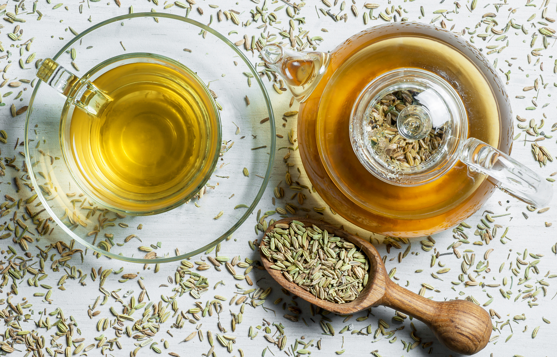 Herbal infusion fennel tea in glass cup and glass tea pot with dried fennel seeds in wooden shovel on white rustic table