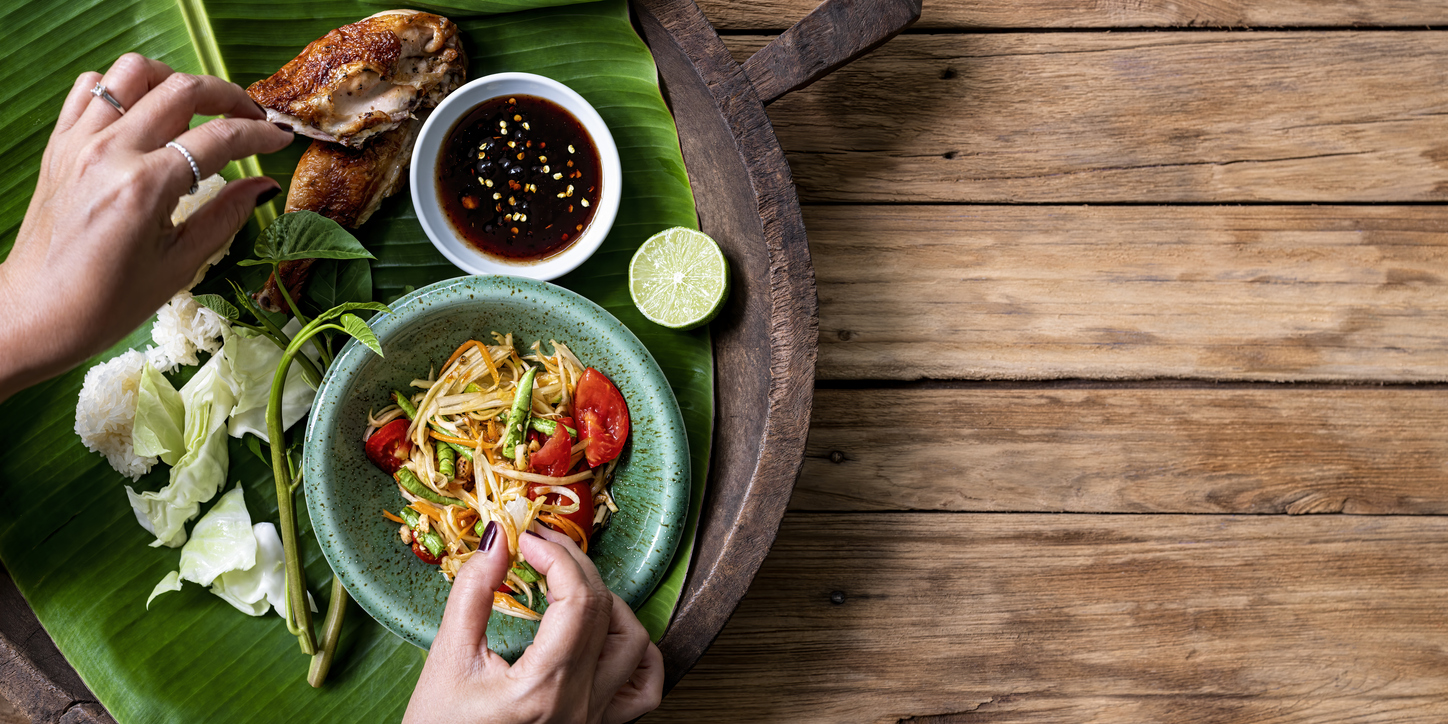 Thai lady eating traditionally with her hands, fresh world famous Som Tam (papaya salad) with BBQ chicken, sticky rice and raw salad vegetables on an old wooden table background.