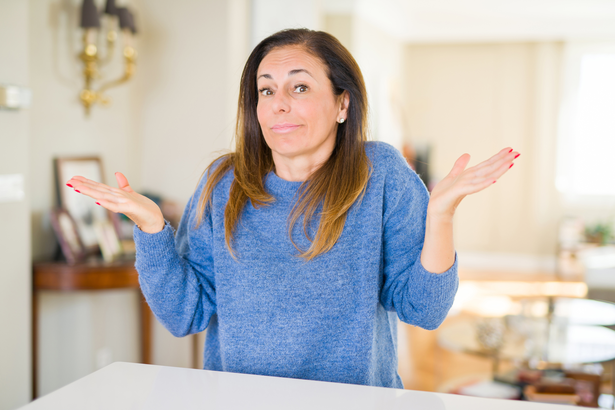 Beautiful middle age woman at home clueless and confused expression with arms and hands raised. Doubt concept.