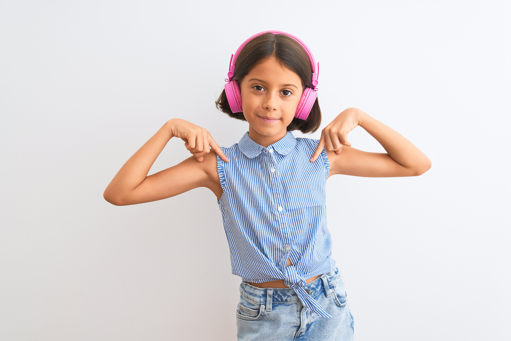 Beautiful child girl listening to music using headphones over isolated white background looking confident with smile on face, pointing oneself with fingers proud and happy.