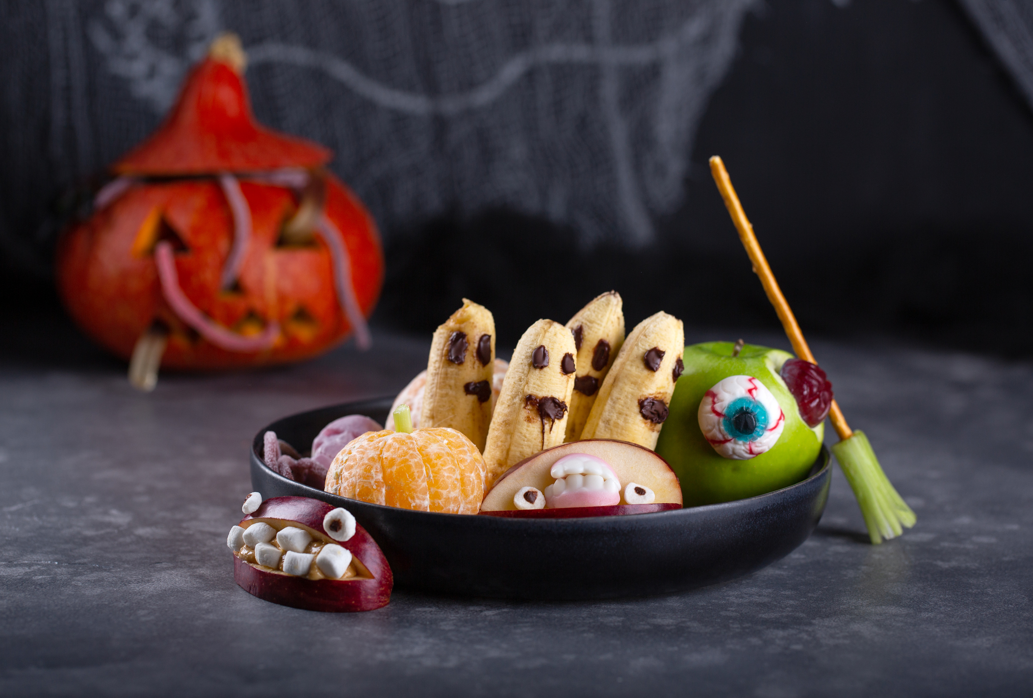 Banana ghosts with chocolate faces, clementine pumpkins and apple monsters on black plate. Fruits in Halloween decoration on dark background.