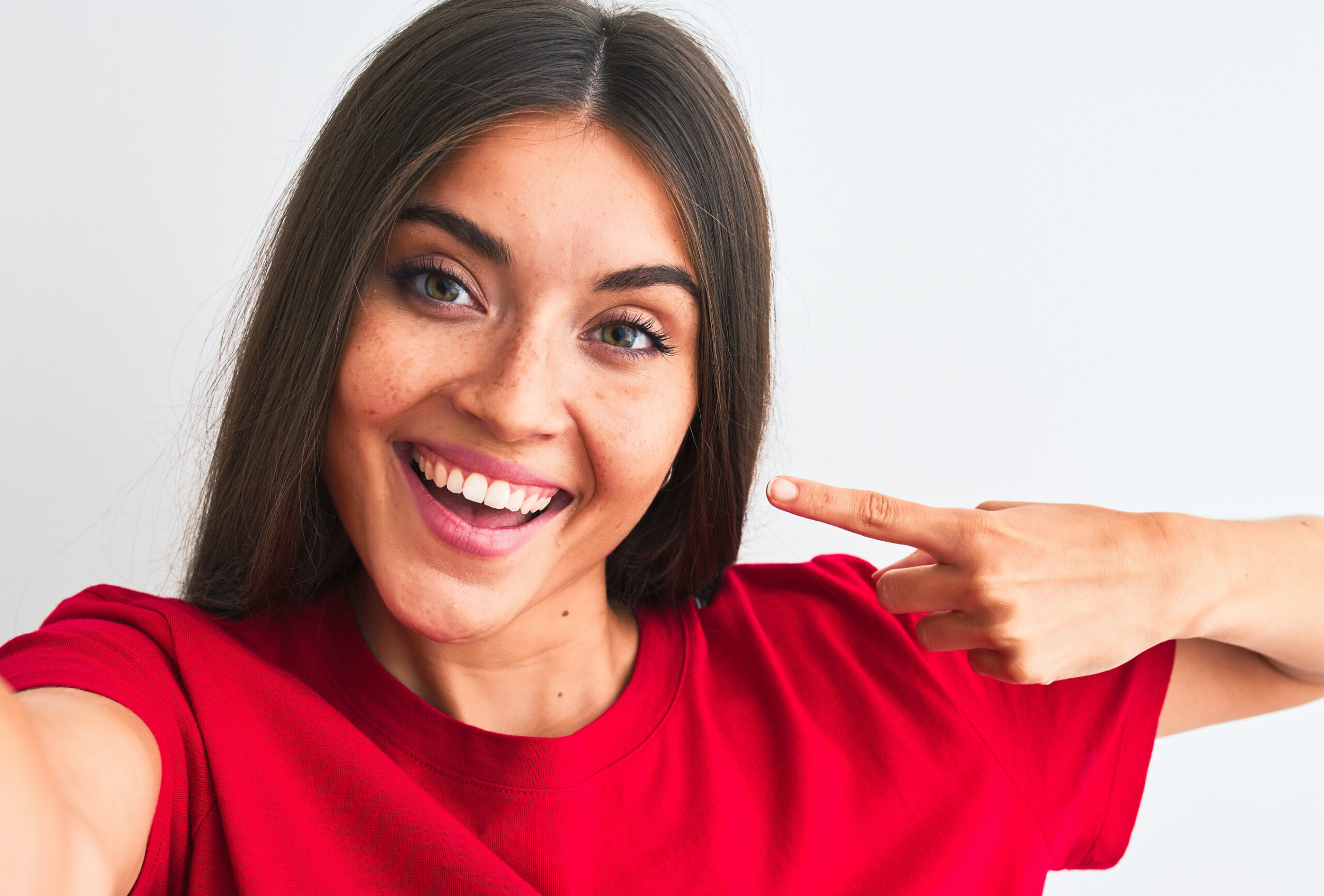 Beautiful woman wearing red t-shirt make selfie by camera over isolated white background very happy pointing with hand and finger