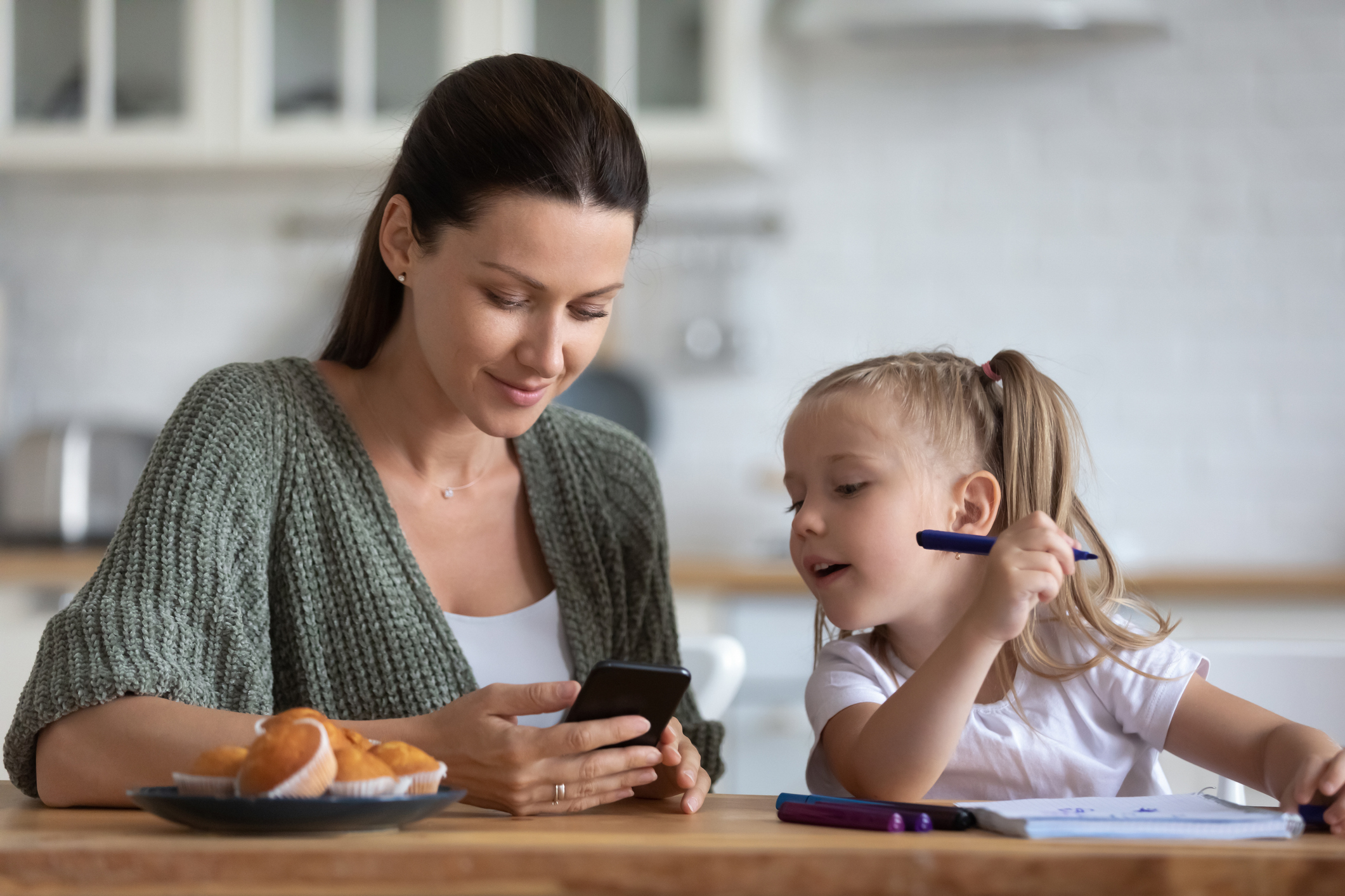 Little daughter distracted from drawing, looking at phone with mother